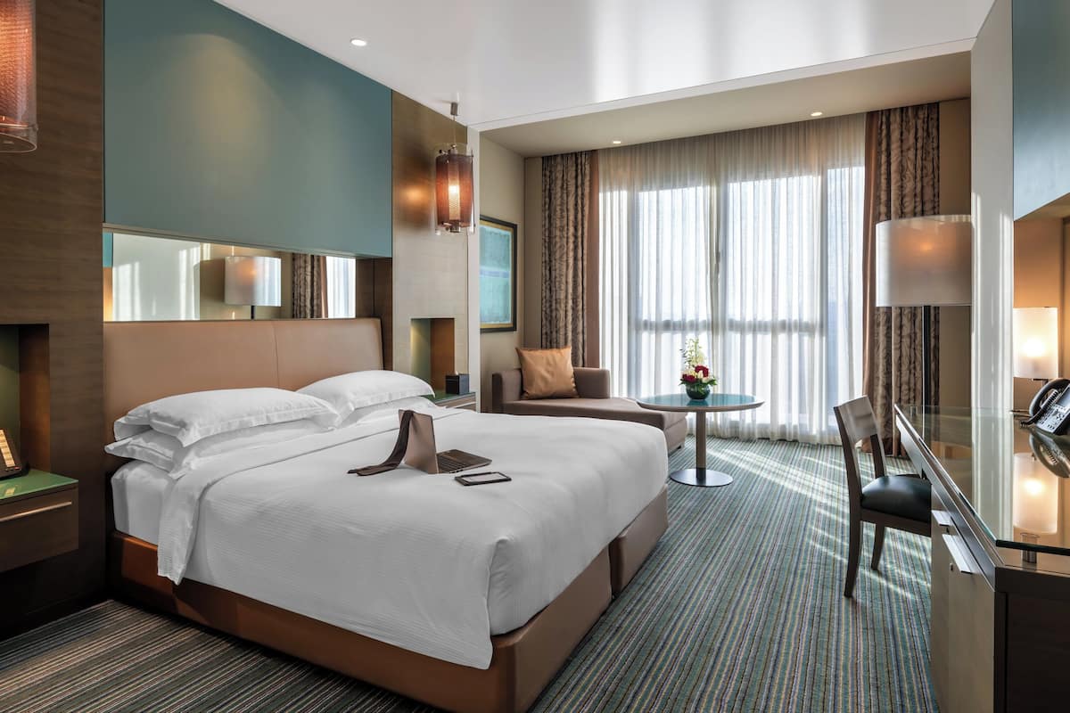 A hotel room with a large bed, a desk with a chair, a small table with a lamp, and a large window with curtains.