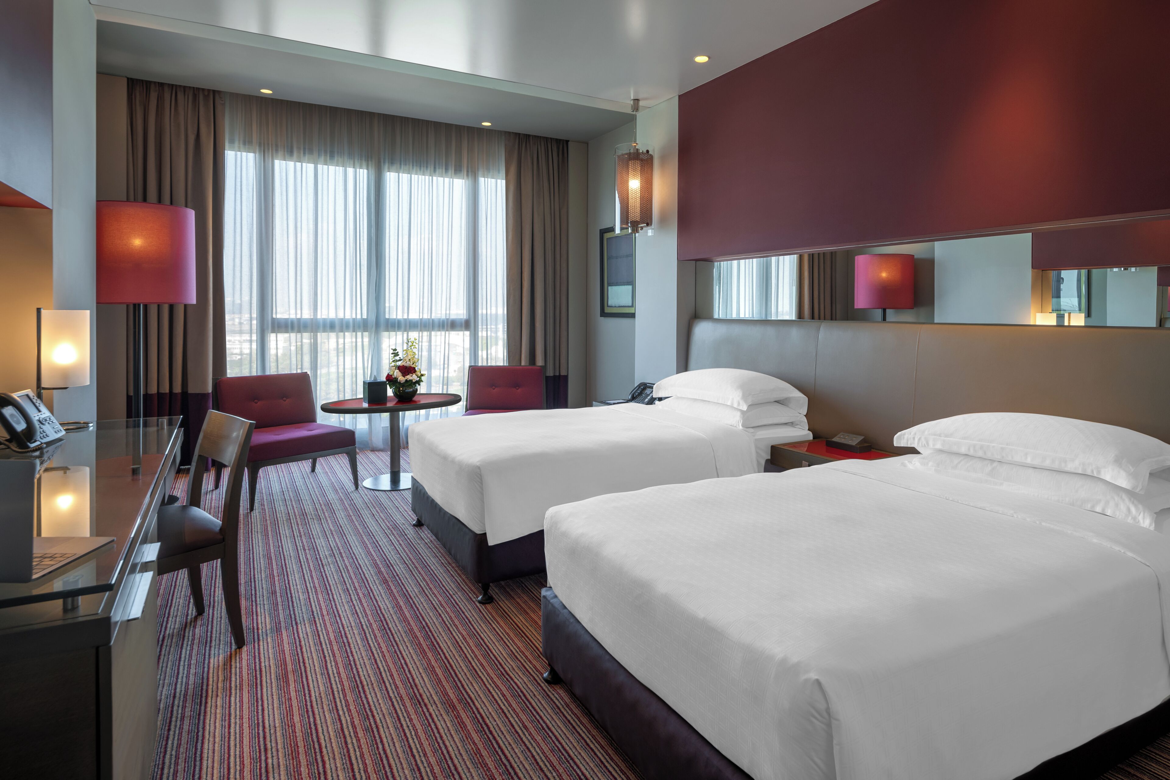 A hotel room with two beds, a desk, a chair, a small table with a vase, and large windows with curtains.