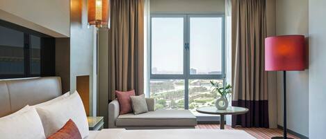 A hotel room with a large bed, a sofa, a round table, and a view of the city through large windows.