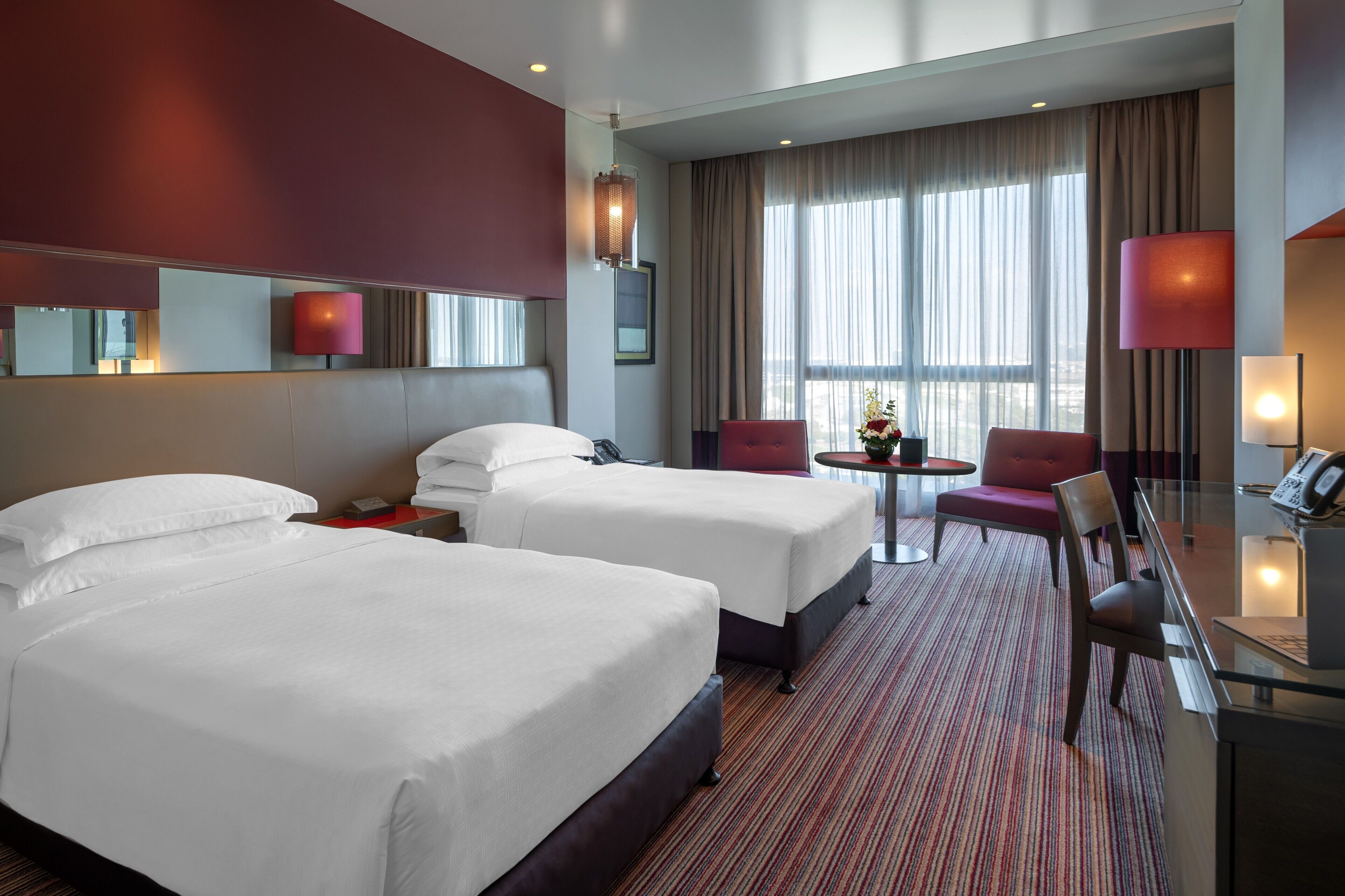 A hotel room with two beds, a desk, a chair, a small table, and a large window with curtains.