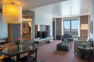A modern hotel room with a flat-screen TV, a sofa, a coffee table, and a dining area with chairs and a glass table.