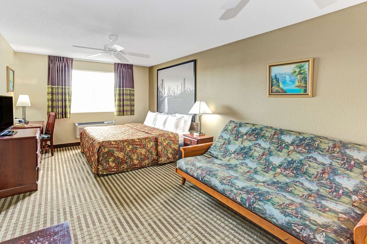A hotel room with a bed, a sofa, a desk, a chair, a ceiling fan, a window with curtains, and a painting on the wall.