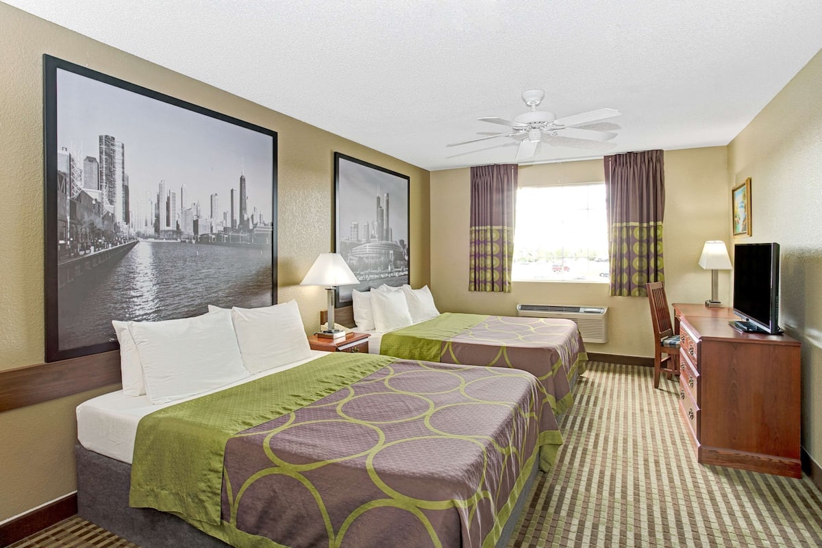 A hotel room with two beds, a desk, a TV, a ceiling fan, and large cityscape pictures on the walls.