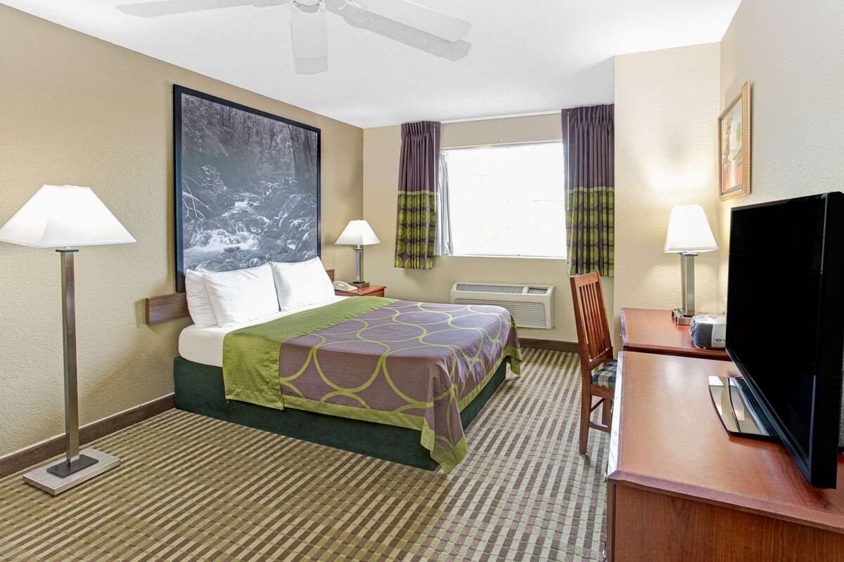 A hotel room with a bed, two bedside lamps, a flat-screen TV, a desk, and a window with curtains.
