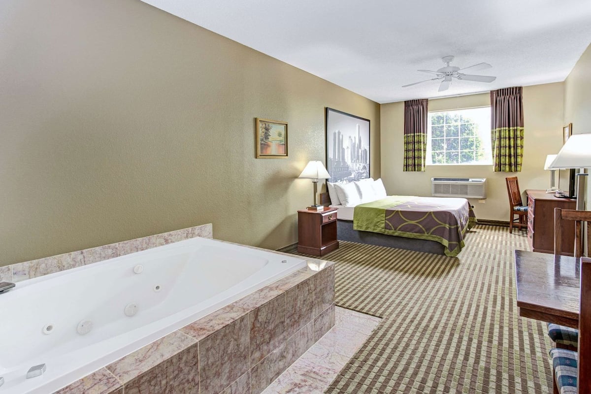A hotel room with a large bathtub, a bed with a green blanket, a desk with a chair, and a window with curtains.