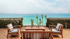 Una terraza de madera con una mesa y cuatro sillas, con vista al mar turquesa y un cielo despejado.