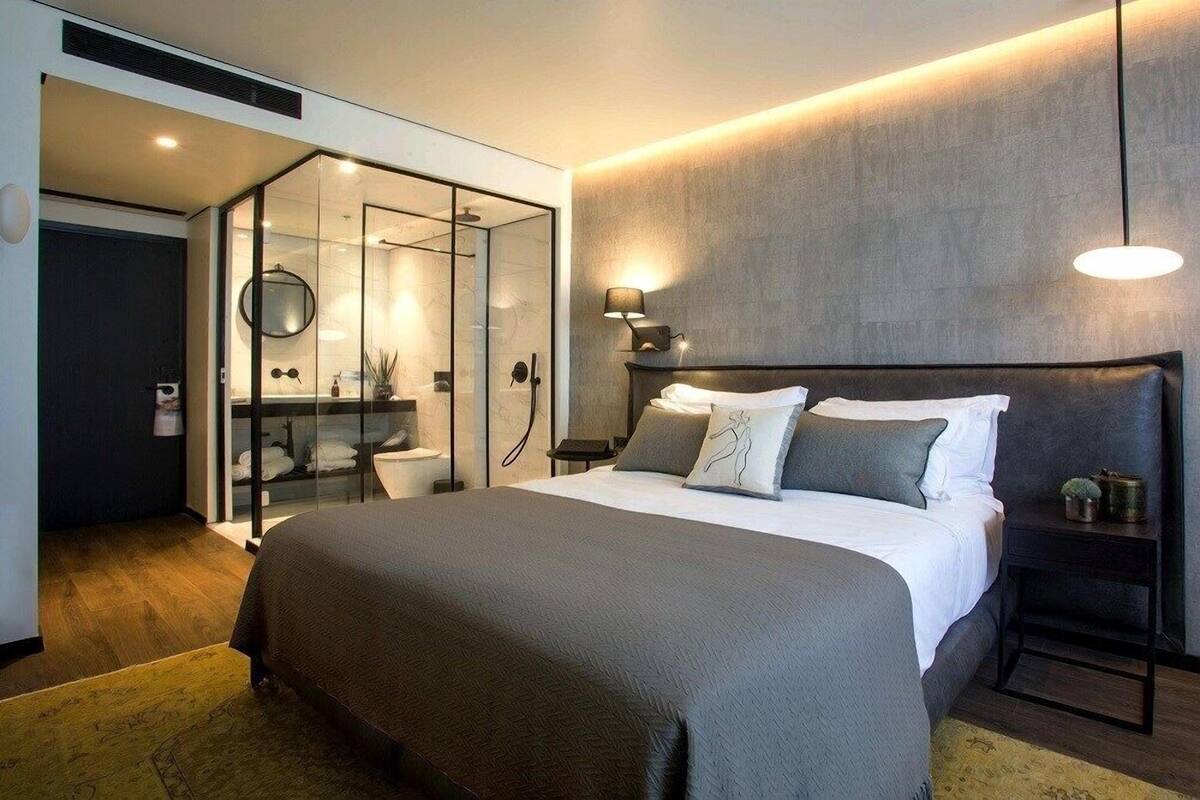 Una habitación de hotel moderna con una cama grande, zona de ducha separada por vidrio y una mesita de noche con lámpara.