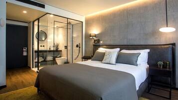 Una habitación de hotel moderna con una cama grande, zona de ducha separada por vidrio y una mesita de noche con lámpara.