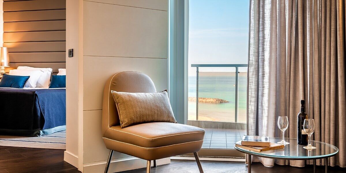 Una habitación de hotel moderna con un gran ventanal, un sillón cómodo, una mesa de vidrio con un libro y copas de vino, y una cama bien hecha.