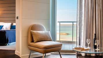 Una habitación de hotel moderna con un gran ventanal, un sillón cómodo, una mesa de vidrio con un libro y copas de vino, y una cama bien hecha.