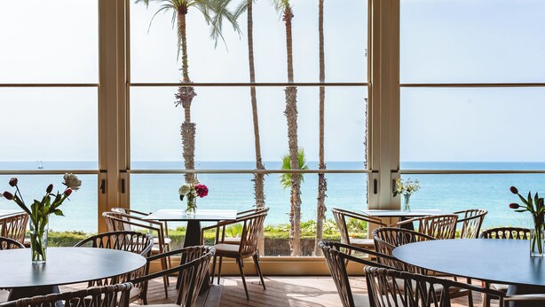 Un restaurante con mesas, sillas, ventanas amplias y vista al océano.