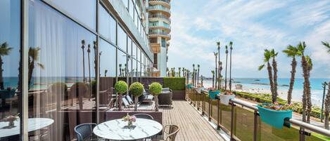 A balcony with outdoor seating, a view of the ocean, and a tall building in the background.