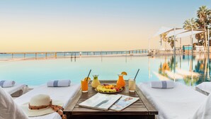 Un área de descanso junto a la piscina con vista al océano, una mesa con toallas, un sombrero y una bandeja de frutas.