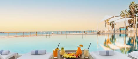 A poolside lounge with a view of the ocean, a table with towels, a hat, and a fruit platter.