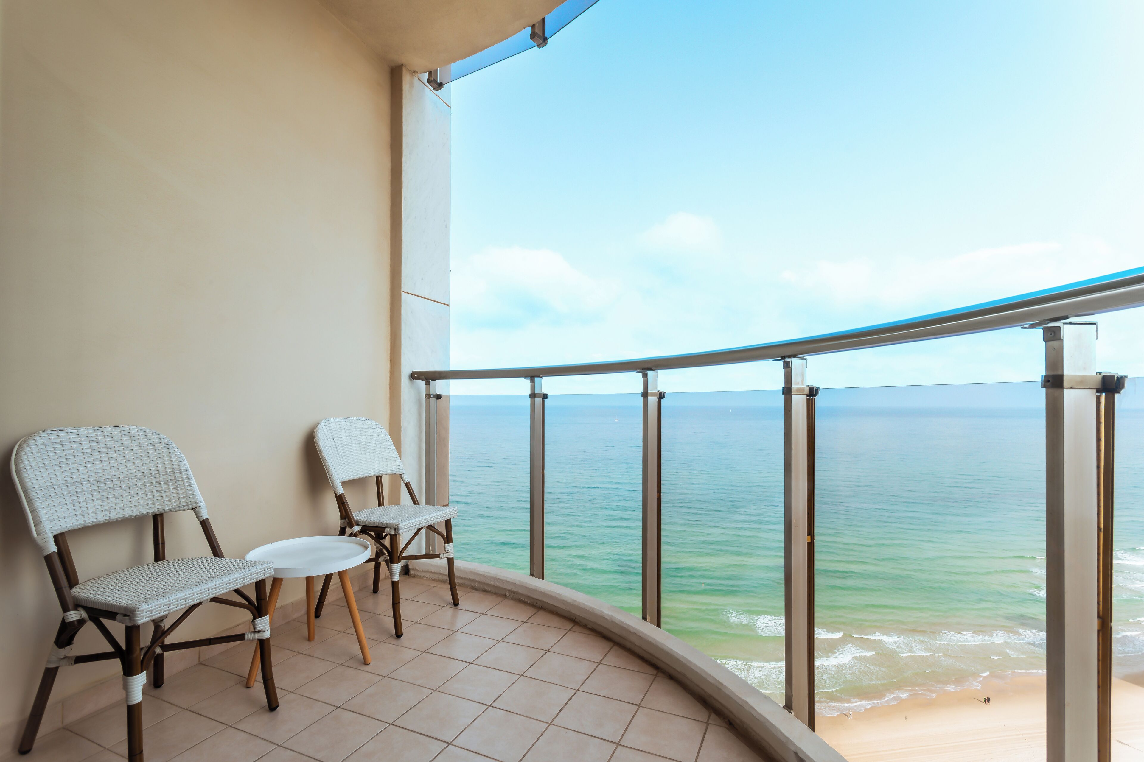Un balcón con dos sillones de mimbre y una mesita, con vistas a una playa de aguas tranquilas y cielo despejado.