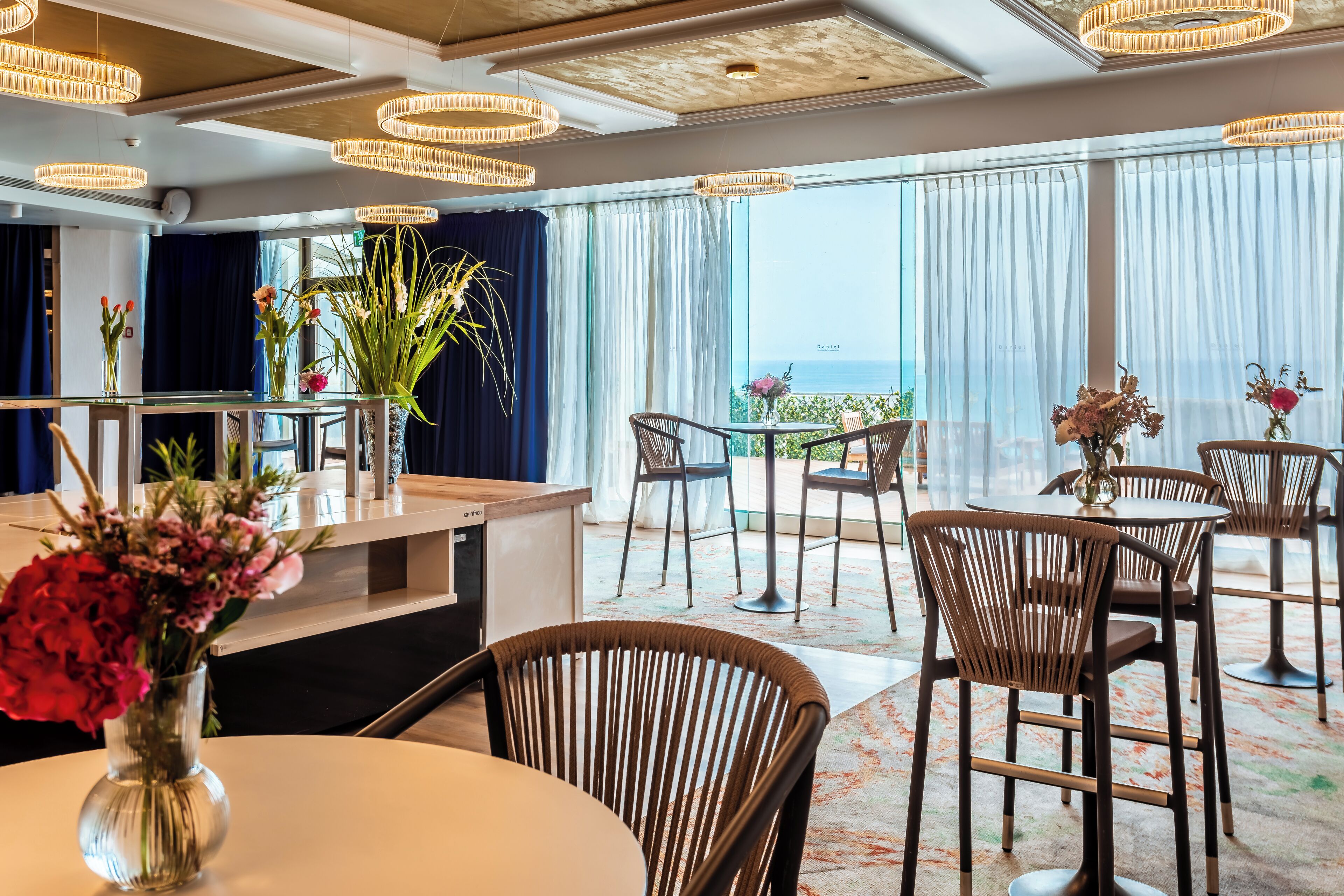 A modern dining area with round tables and wicker chairs, large windows with curtains, and a view of the sea.
