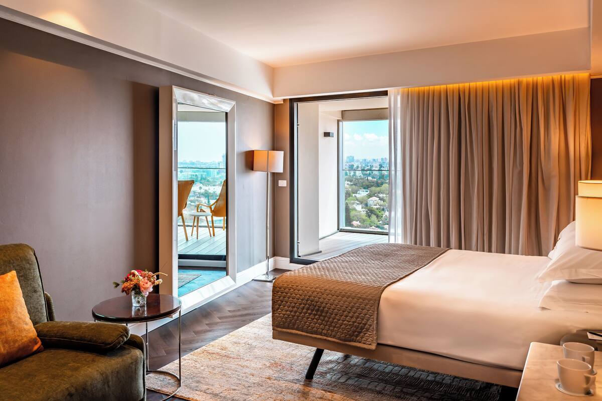 Una habitación de hotel moderna con una cama grande, un rincón de descanso con una silla, una mesita con un jarrón de flores y vistas a la ciudad a través de amplios ventanales.