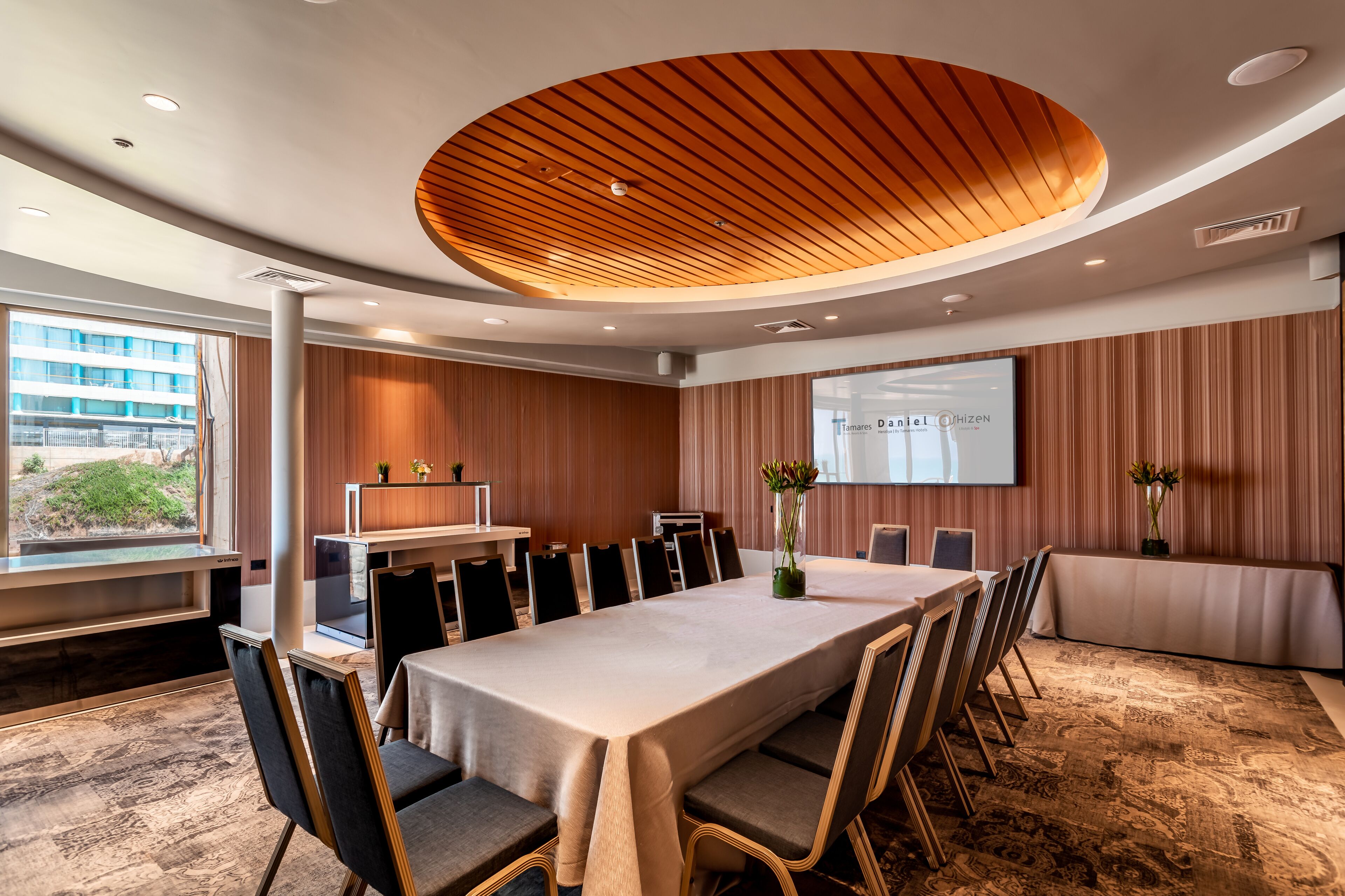 Un salón de conferencias con una larga mesa, sillas, una pantalla de proyector y techo de madera.