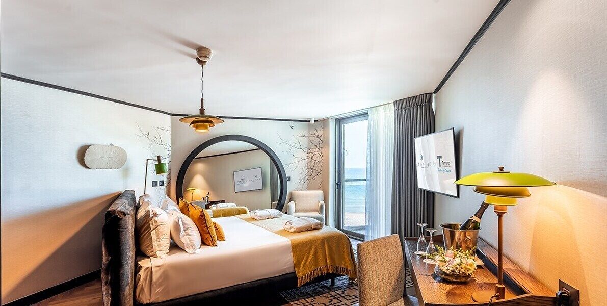 Een hotelkamer met een bed, een bureau met een lamp, een stoel, een raam met gordijnen en uitzicht op zee.