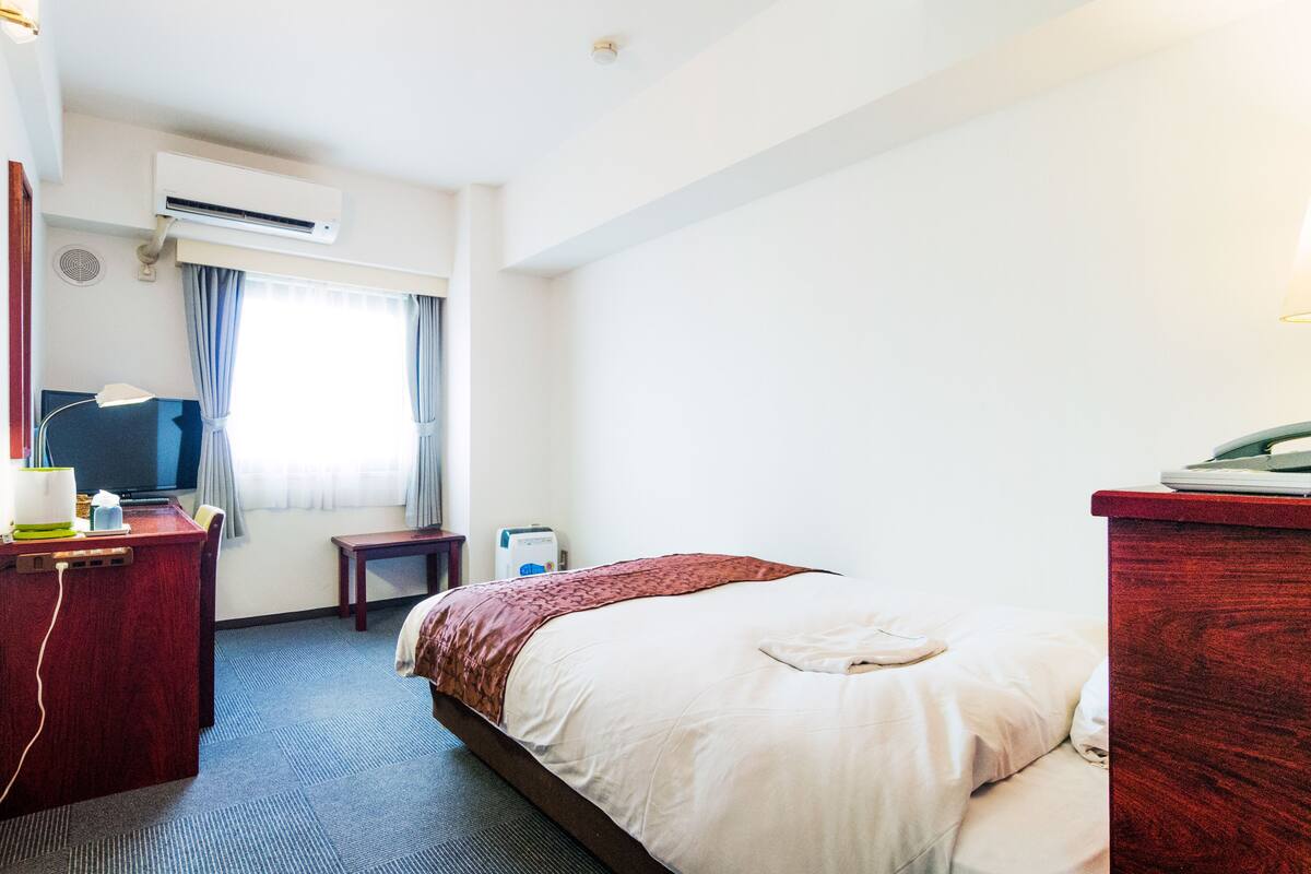 Une chambre d’hôtel équipée d’un lit, d’un bureau, d’un climatiseur, d’une fenêtre avec des rideaux et d’une table de chevet.