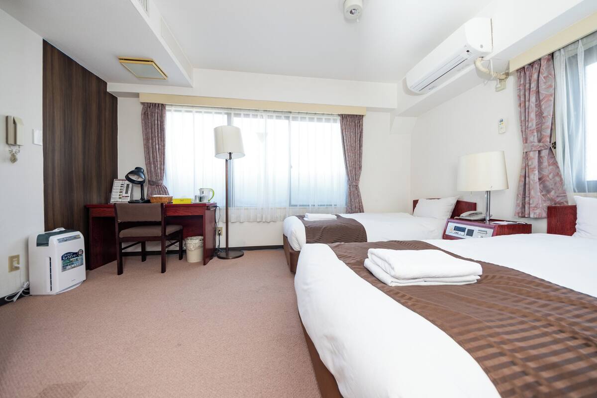 Une chambre d’hôtel avec deux lits, un bureau avec une chaise, une lampe, un climatiseur et une table de chevet avec une lampe.