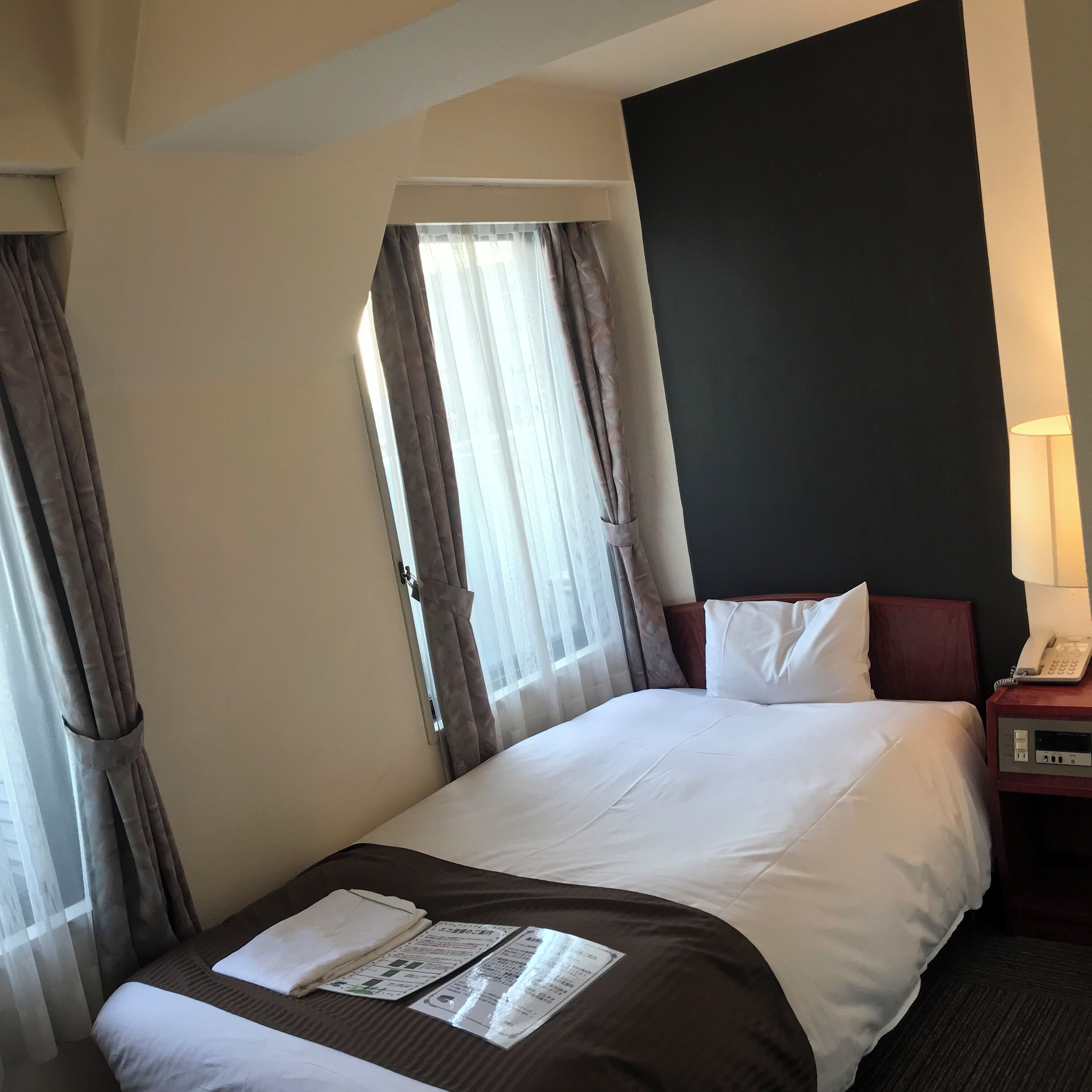 Una camera d'albergo con un letto, un comodino con una lampada e una finestra con tende.