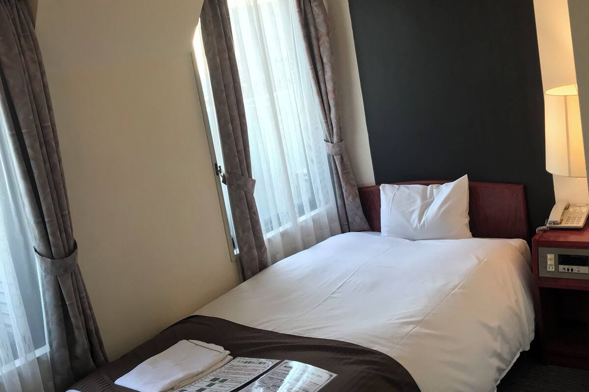 Une chambre d’hôtel avec un lit, une table de chevet avec une lampe et une fenêtre avec des rideaux.