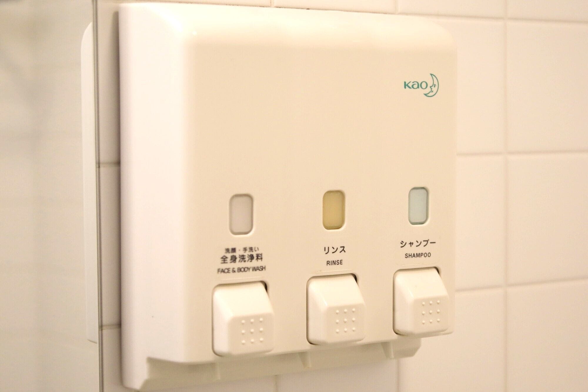 Un distributeur blanc, fixé au mur, doté de trois boutons indiquant les fonctions suivantes : lavage du visage et du corps, rinçage et application de shampooing.
