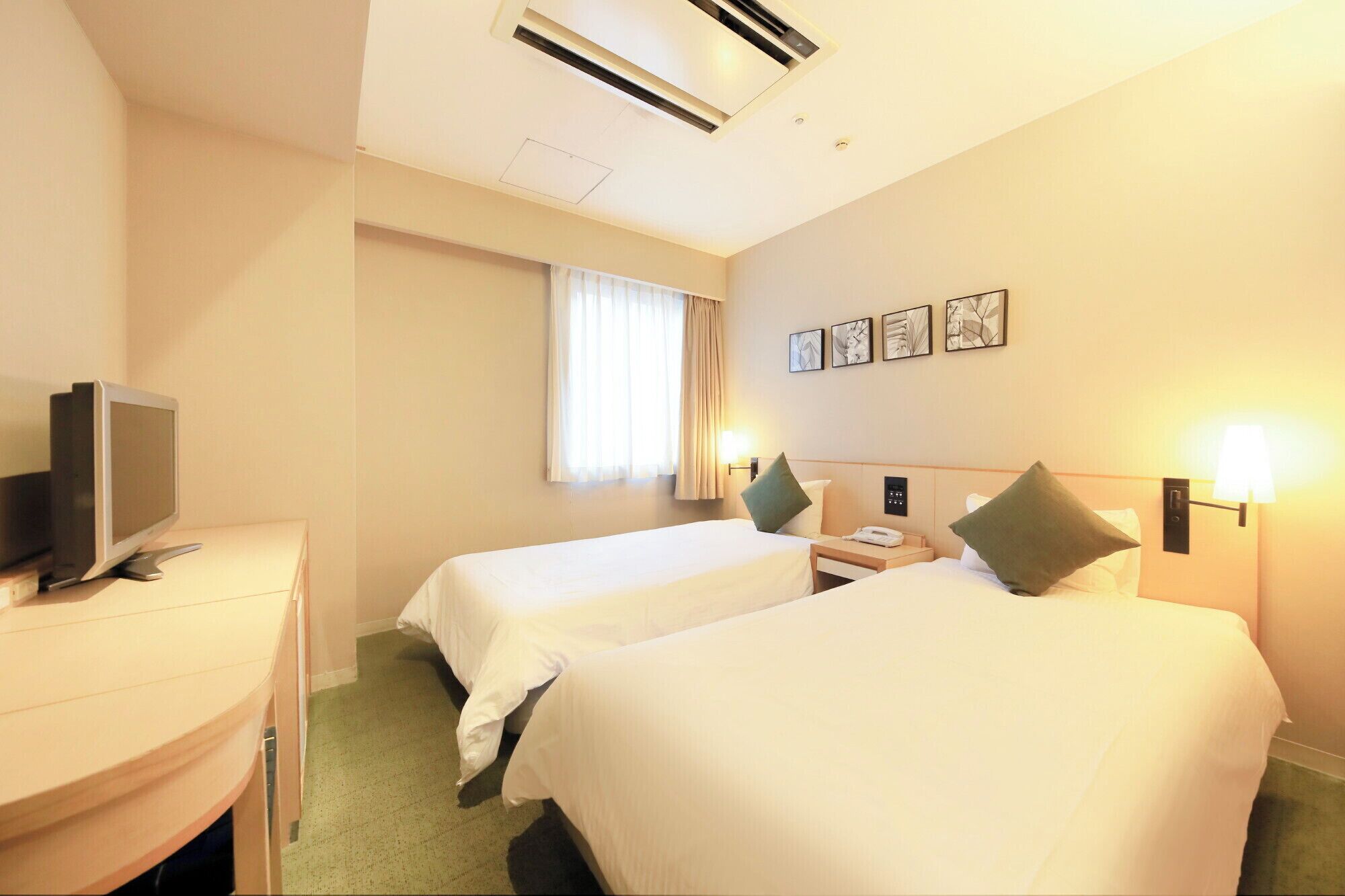 Quarto de hotel com duas camas, uma escrivaninha com televisão, uma janela com cortinas e luminárias fixadas na parede.