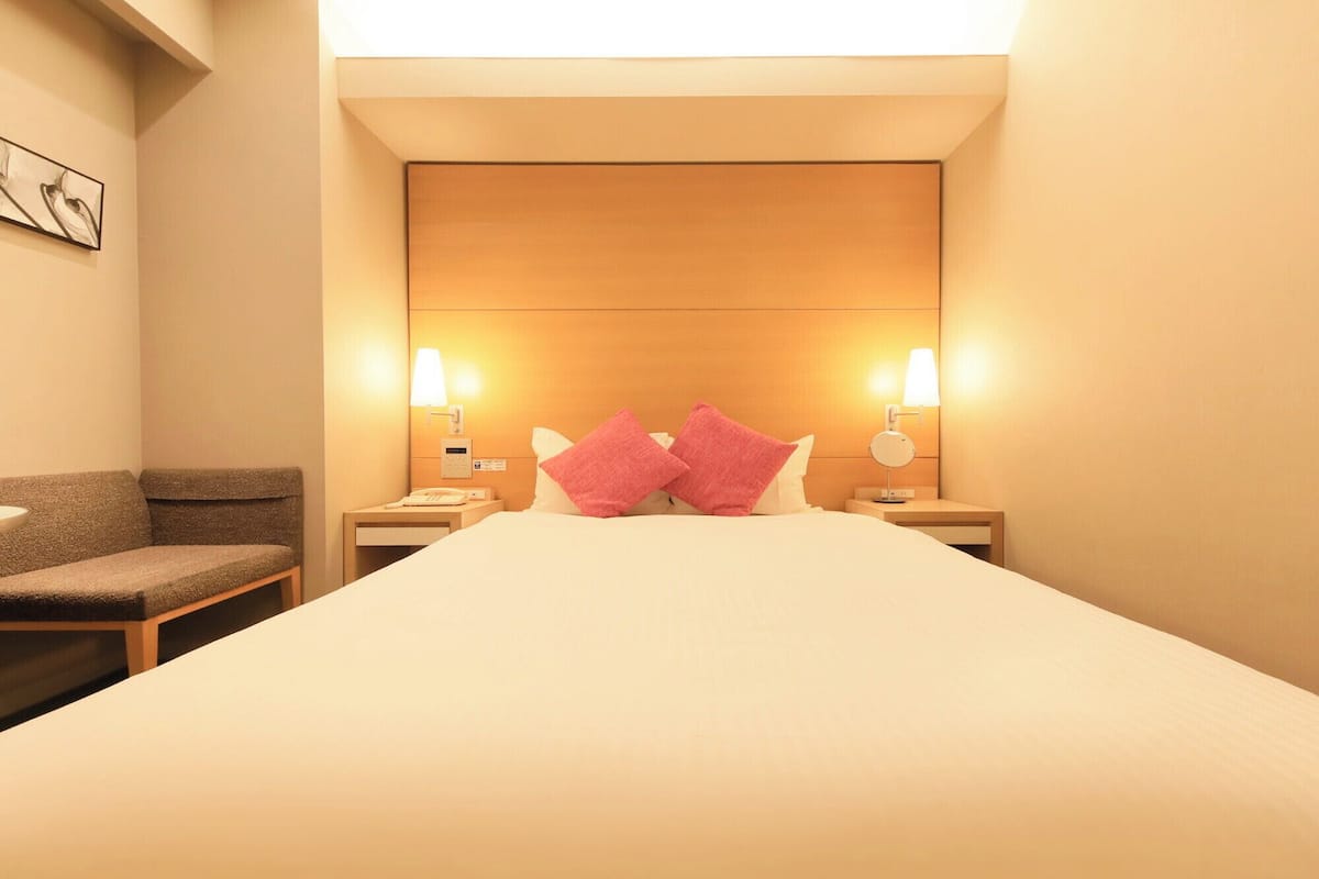 A hotel room with a large bed, two bedside lamps, a nightstand with a phone, and a chair.