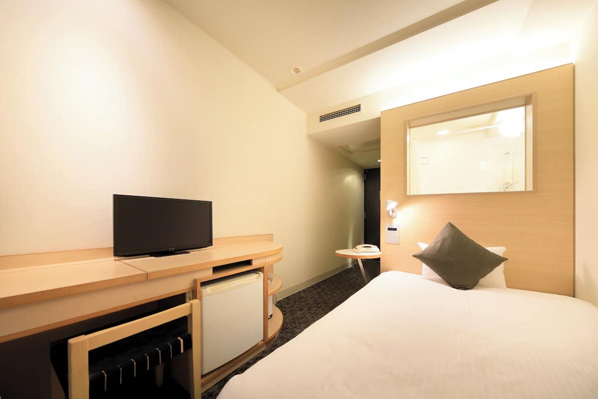 A hotel room with a bed, a desk with a television, a small table with a phone, and a mirror on the wall.