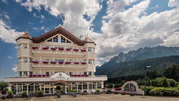A multi-story hotel with a classic European architectural style, featuring balconies with flower boxes, a prominent entrance, and a view of mountains in the background.