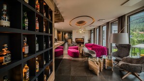Een modern interieur met een lange plank vol drankflessen, een gezellige zithoek met een roze poef, en uitzicht op een bergachtig landschap door grote ramen.