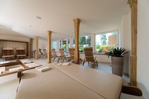 A spa room with wooden chairs, a massage table, and large windows offering a view of greenery. - Park Hotel Bellevue (Dobbiaco)
