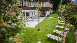 Een modern gebouw met een verzorgd gazon, een terras met ligbedden en een kleine boom in het midden.