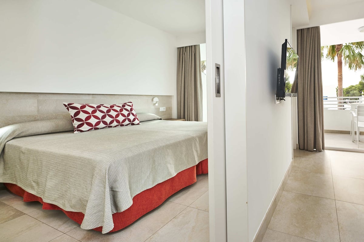 A hotel room with a bed, a TV, a balcony with a table and chairs, and a view of palm trees.