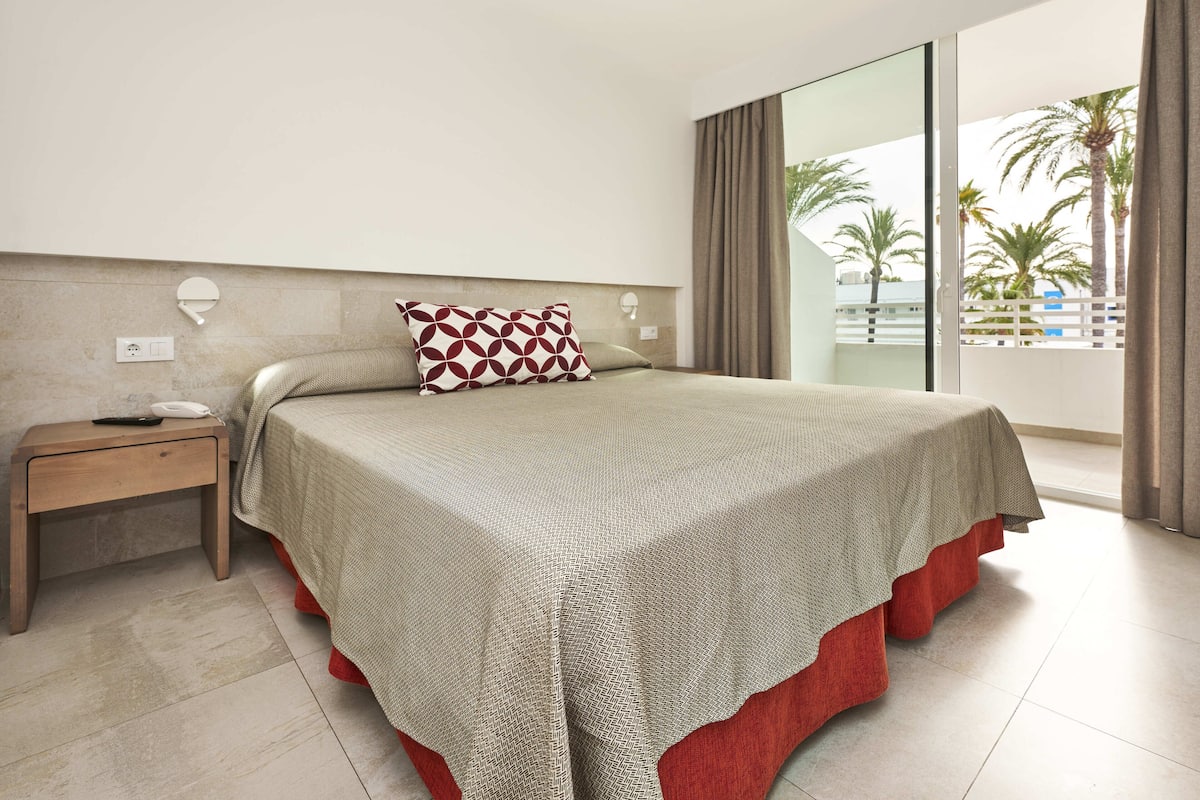A modern hotel room with a large bed, a bedside table with a phone and a remote, and a view of palm trees through a large window.
