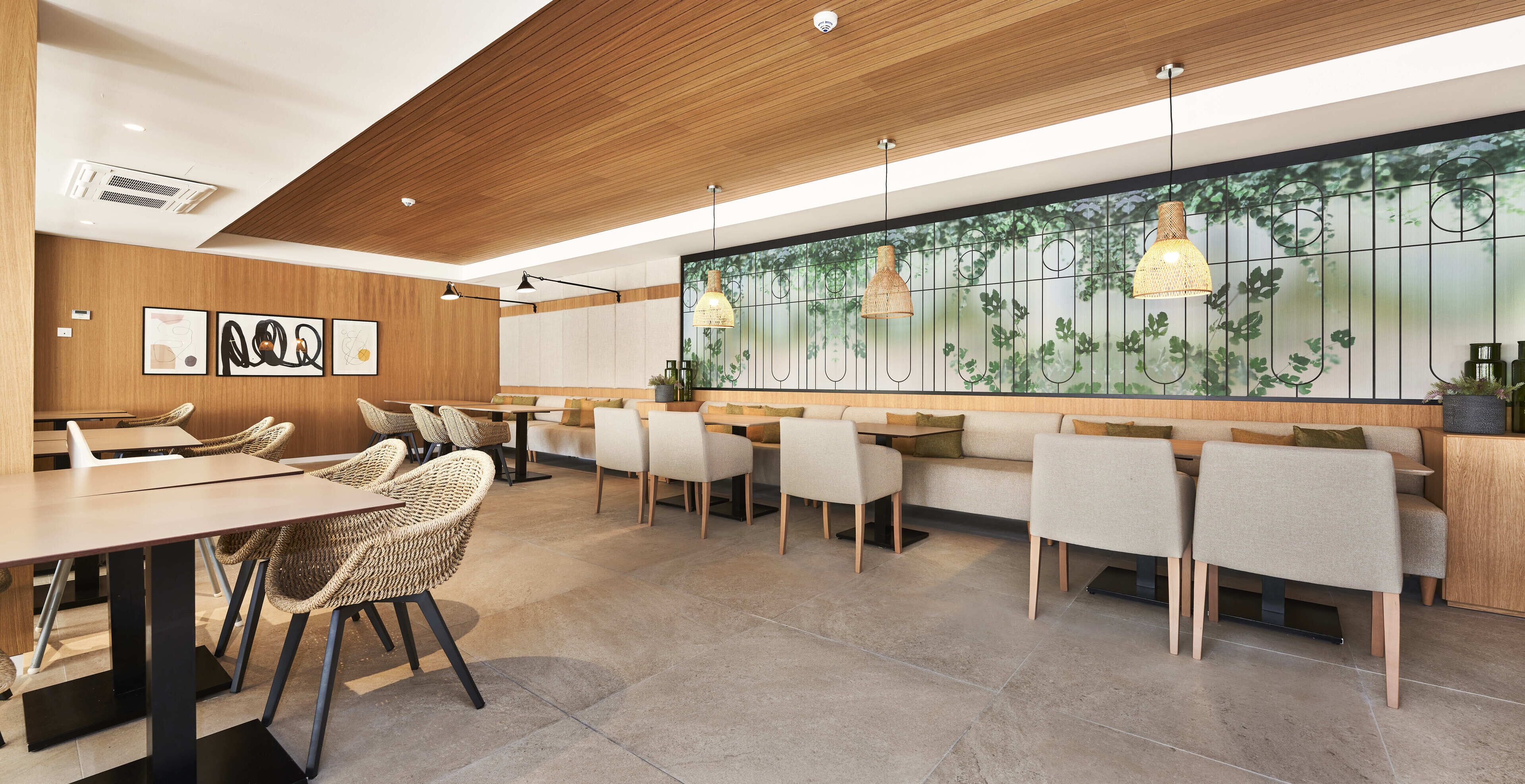 A modern restaurant with wooden ceiling, tiled floor, and a large mural of trees. There are several tables and chairs, some with cushions.