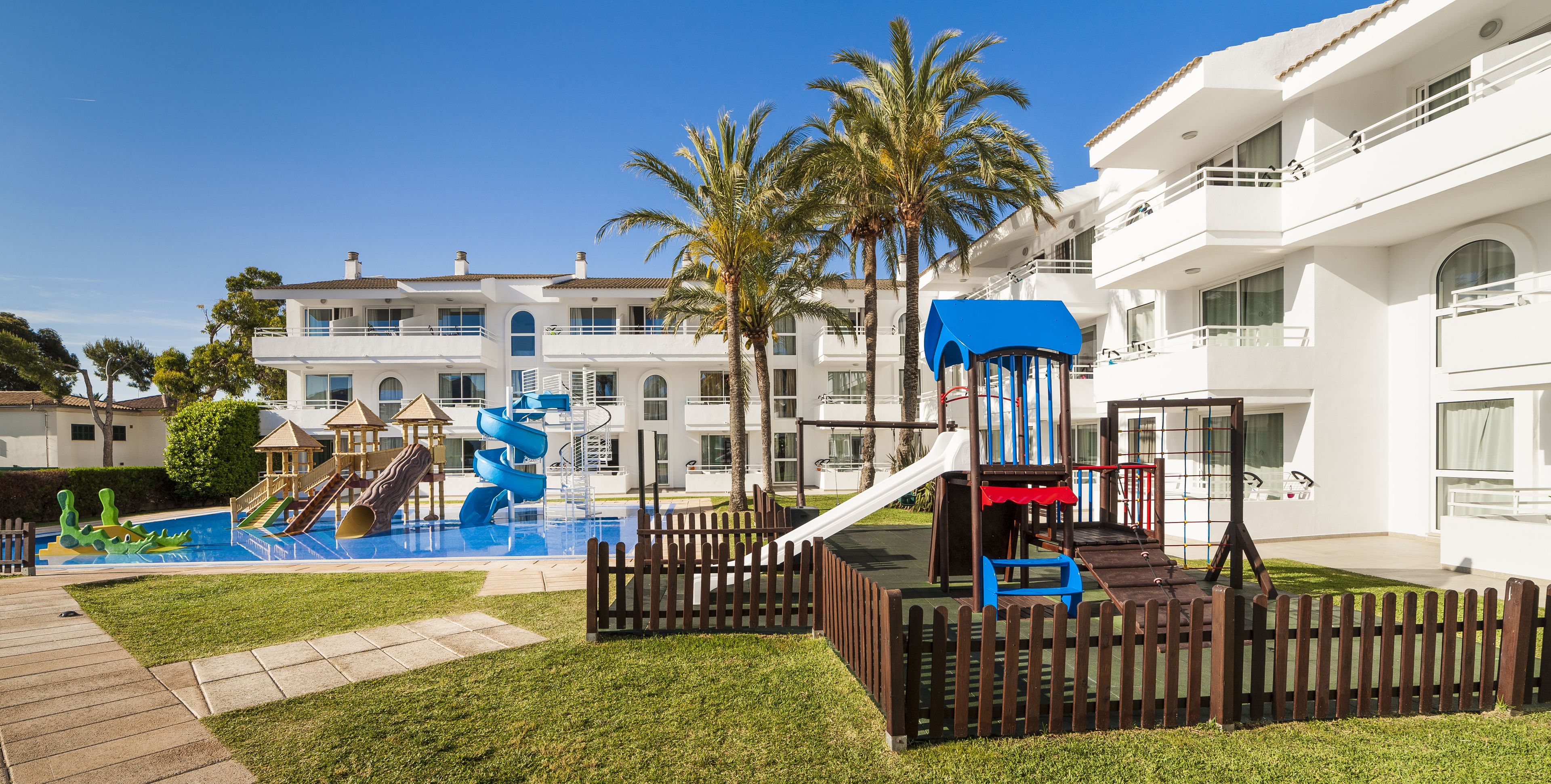 A children's play area with slides and a pool, surrounded by a wooden fence, and a white building with balconies in the background.