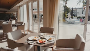 A dining area with a table set for two, a view of the pool area, and a modern interior design. - Hoposa Hotel & Apartments Villaconcha (Pollenca)