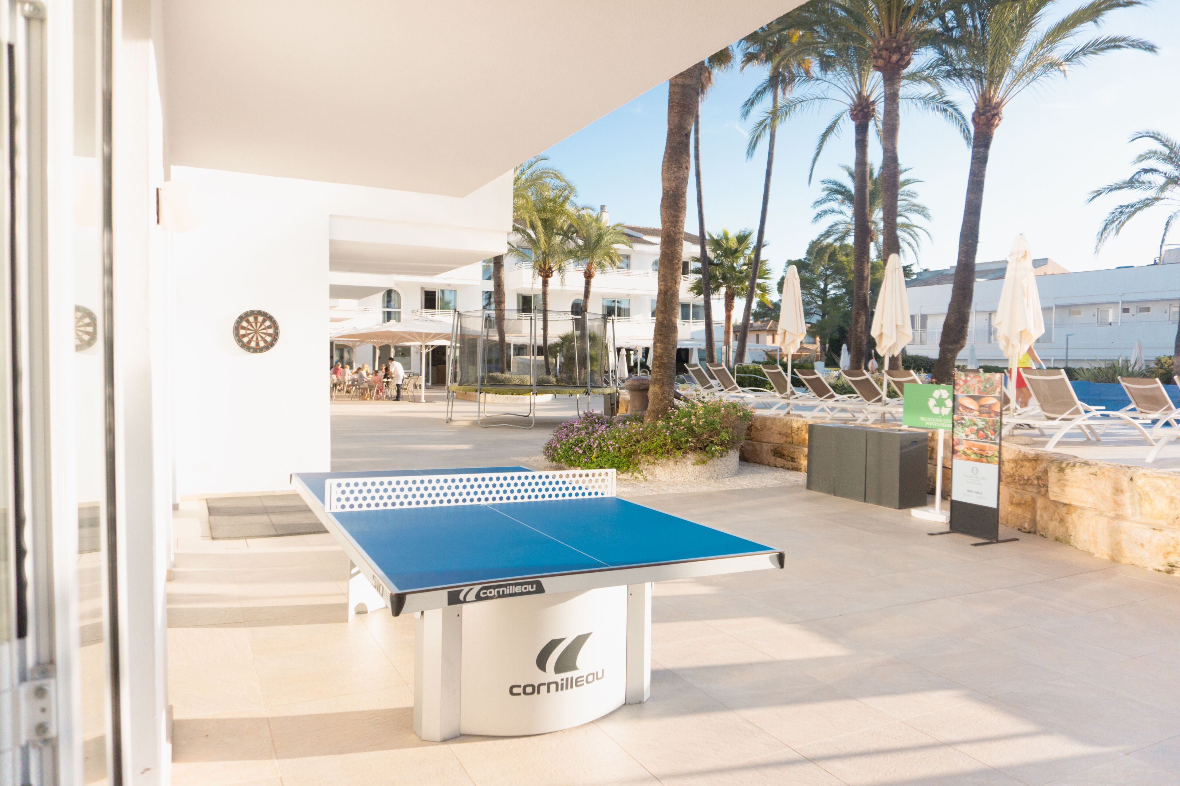 An outdoor ping pong table with a white and blue surface, surrounded by lounge chairs and palm trees.