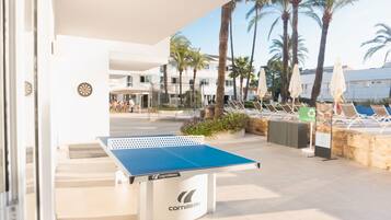 An outdoor ping pong table with a white and blue surface, surrounded by lounge chairs and palm trees.