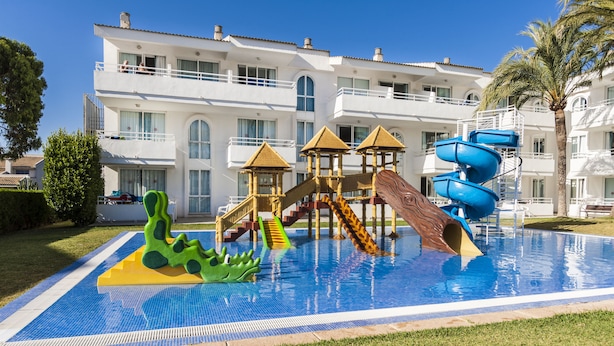 A swimming pool with slides and a play structure, surrounded by a well-maintained lawn and a large building in the background. - Hoposa Hotel & Apartments Villaconcha (Pollenca)