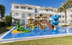 Una piscina con toboganes y una estructura de juegos, rodeada por un césped cuidado y un edificio grande al fondo.