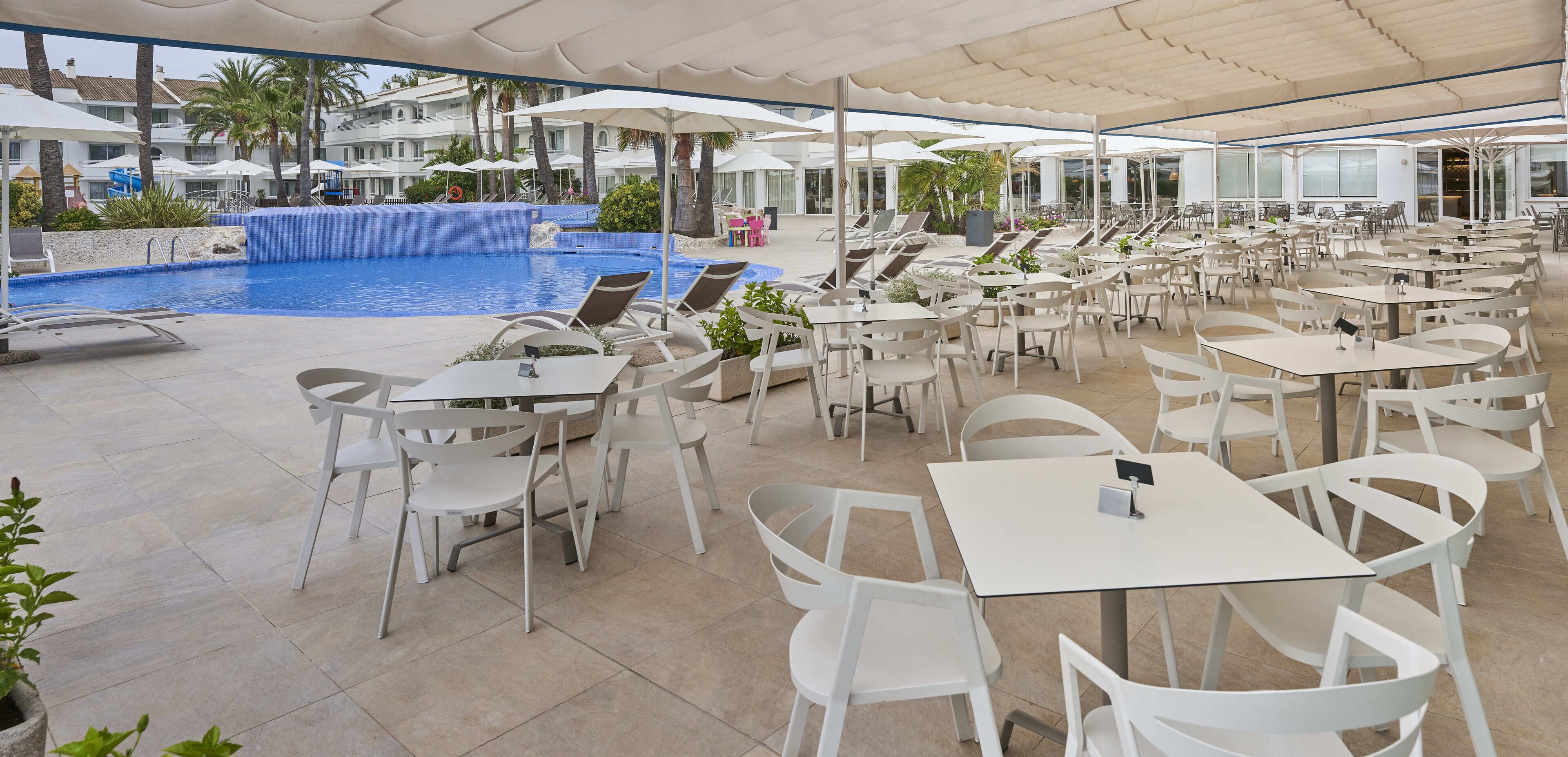 Un espace aménagé au bord de la piscine, avec des chaises et des tables blanches, une piscine et des parasols.