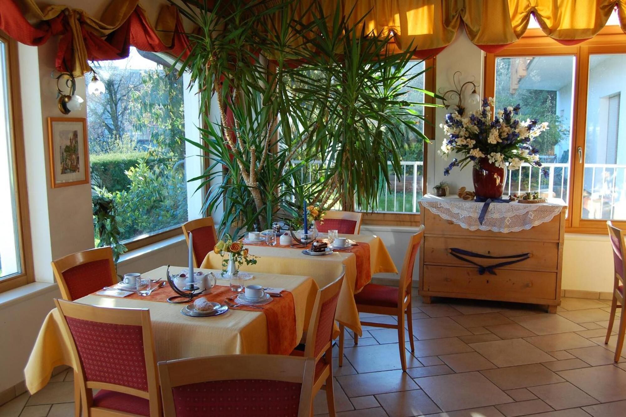 Zona pranzo con tavolo apparecchiato per quattro, circondata da sedie, una grande pianta in vaso e un settimino con un vaso di fiori.