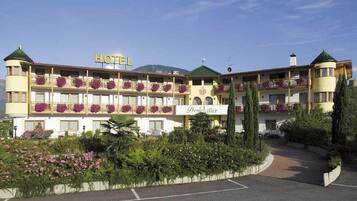 Classic hotel with a central entrance and balcony overlooking a well-maintained garden and mountainous landscape.