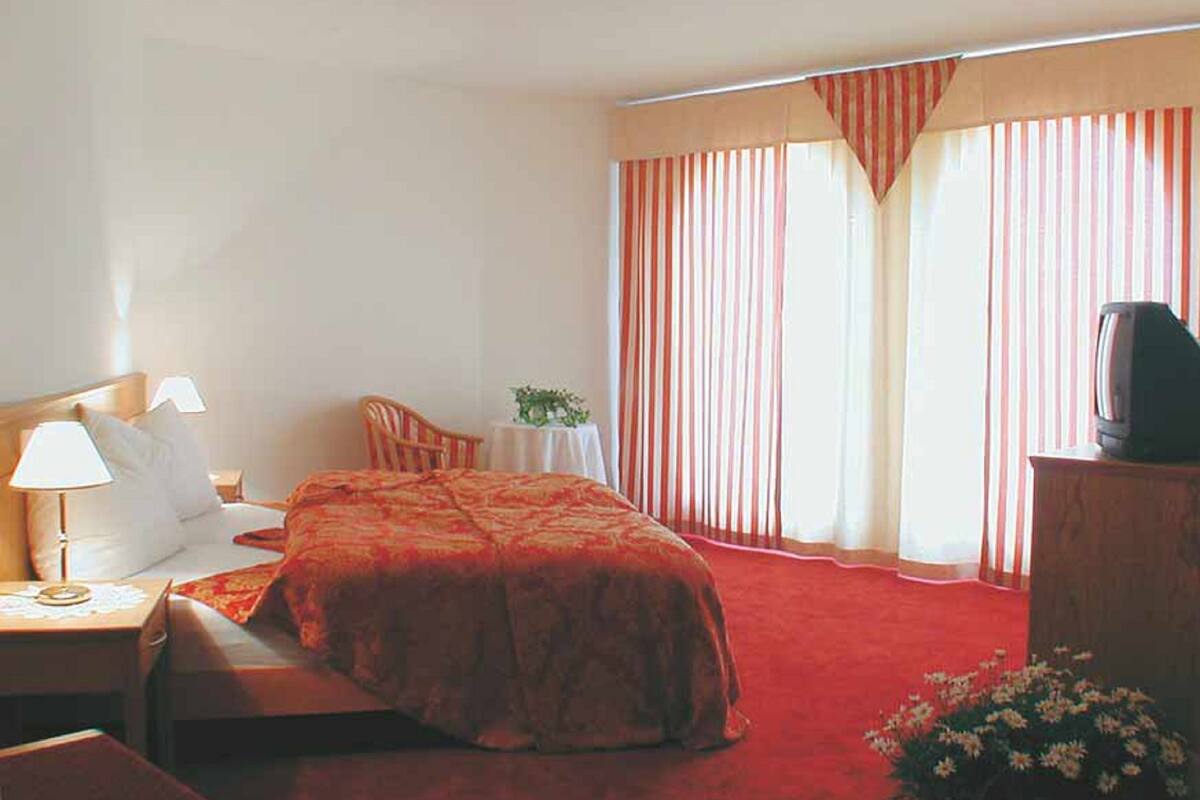 A hotel room with a bed, a desk with a lamp, a chair, a small table with a plant, and a television on a cabinet.