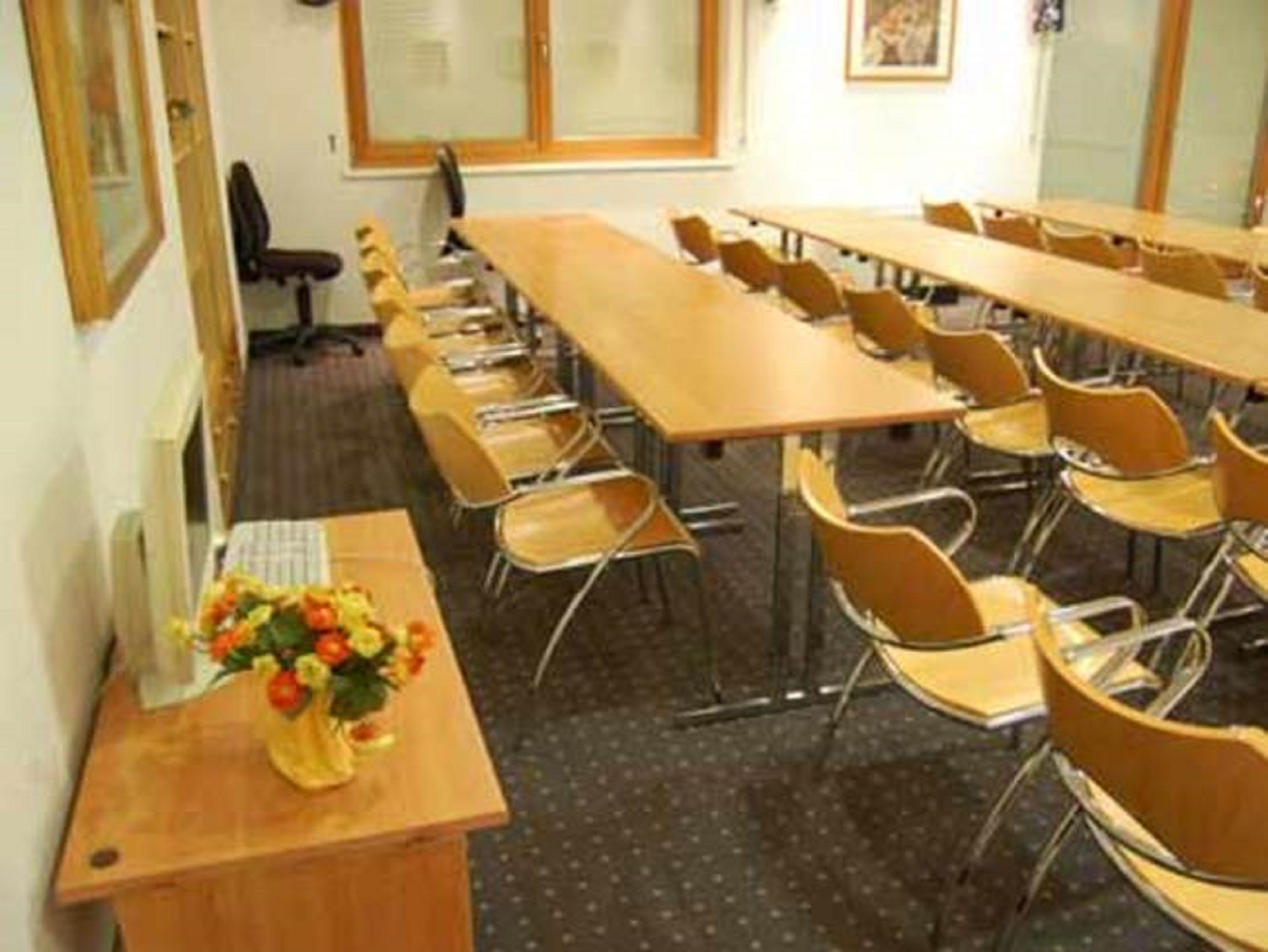 Ein Konferenzraum mit langen Tischen und Stühlen, ein Holztisch mit Blumen in einer Vase und ein Fenster mit Jalousien.