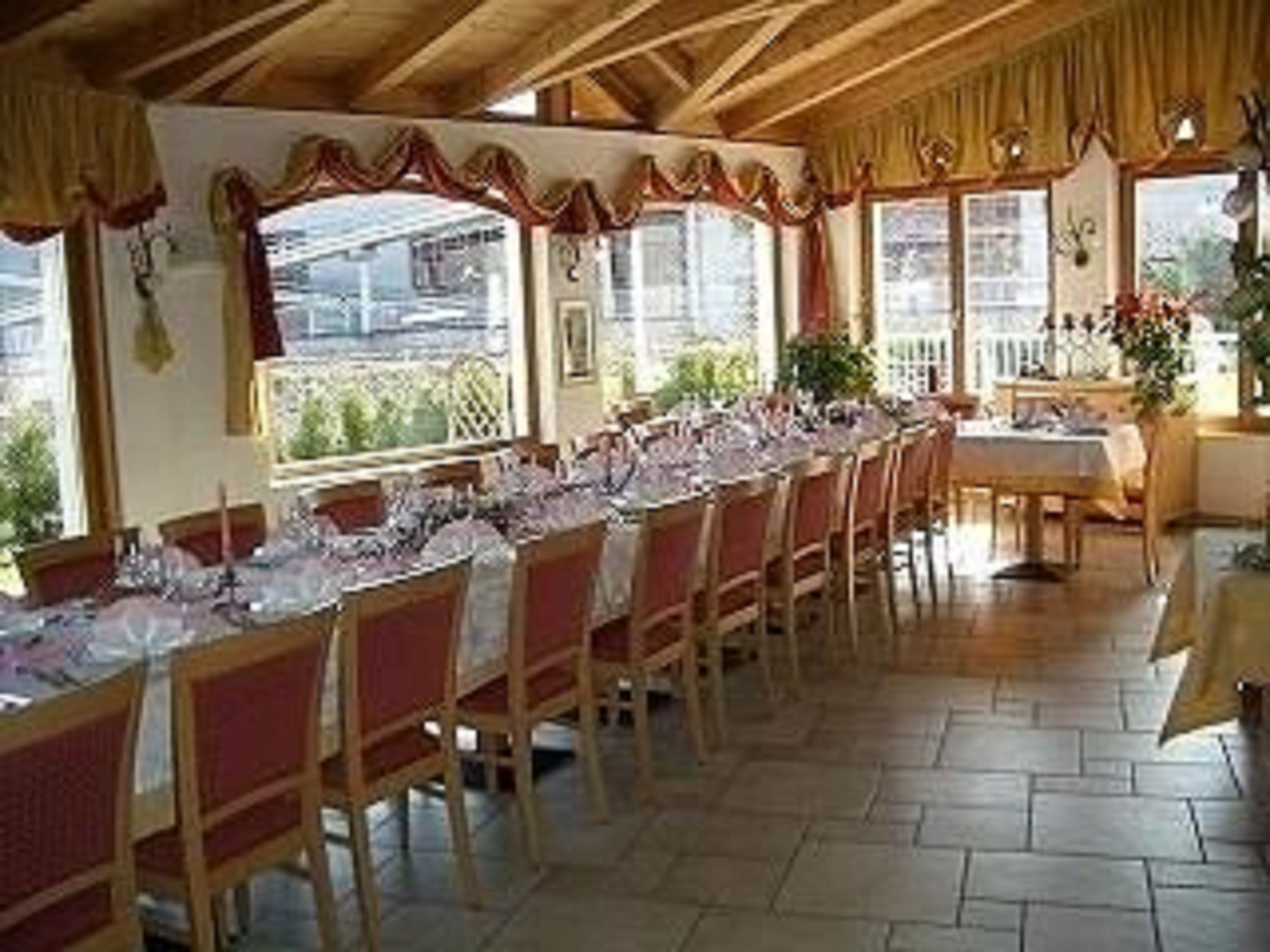 Ein Essbereich mit einem langen Tisch, der für eine Mahlzeit gedeckt ist, Holzstühlen und großen Fenstern mit Vorhängen.